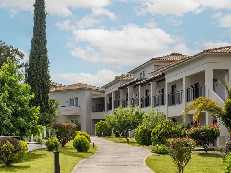 Großes Wohnhaus mit mehreren Stockwerken, umgeben von grünen Büschen, Bäumen und einem geschwungenen Weg.