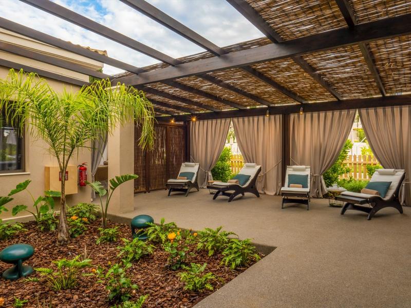 Überdachte Terrasse mit Pflanzenbeet, Sonnenliegen und braunen Vorhängen an Holz-Glasdach.