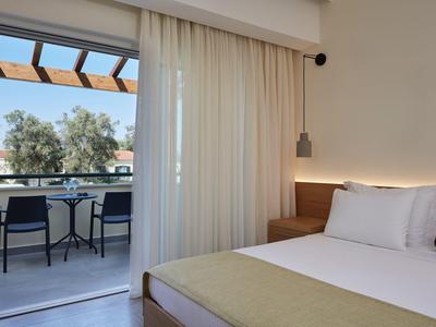 Helles Hotelzimmer mit Bett und Zugang zu Balkon mit Tisch und zwei Stühlen.