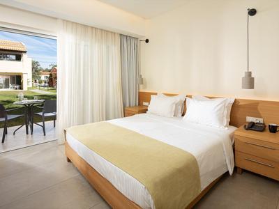 Helles Hotelzimmer mit großem Bett, Nachttisch, und Blick auf Terrasse mit Garten