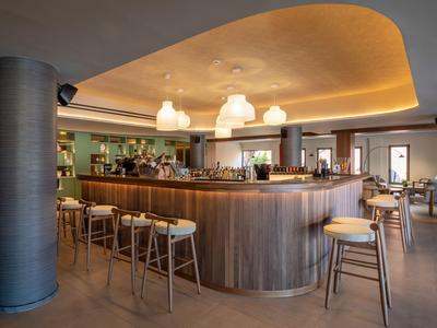 Innenansicht einer modernen Hotelbar mit Holztheke und Barhockern unter warm beleuchtetem Deckenelement.