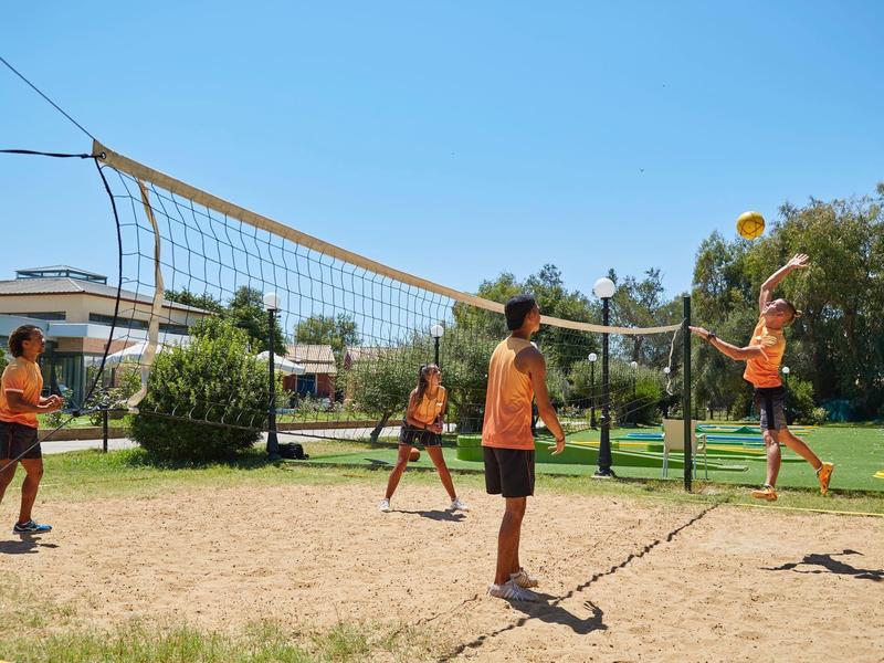 Kinder spielen Volleyball auf einem Sandplatz bei sonnigem Wetter in einem Park.