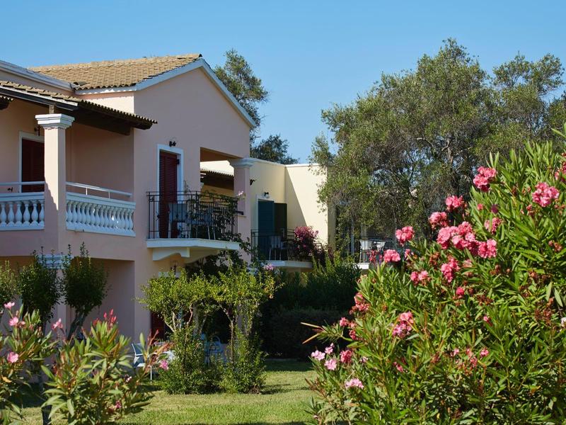 Haus mit Balkon und grüner Gartenanlage bei blauem Himmel und blühenden Sträuchern