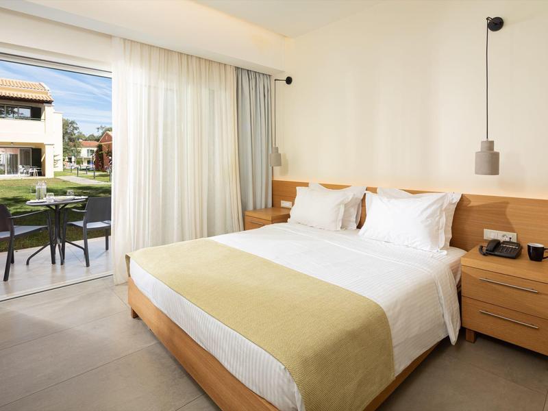 Helles Hotelzimmer mit großem Bett, Nachttisch, und Blick auf Terrasse mit Garten