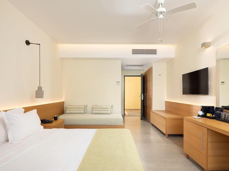 Helles Hotelzimmer mit Doppelbett, Sofa, Fernseher an der Wand und Schreibtisch mit Kaffeezubehör.