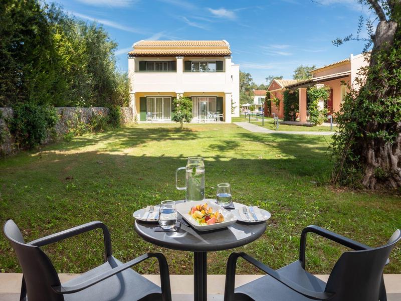 Moderner Tisch mit Frühstück vor einem weißen Haus mit Garten und Bäumen im Hintergrund.