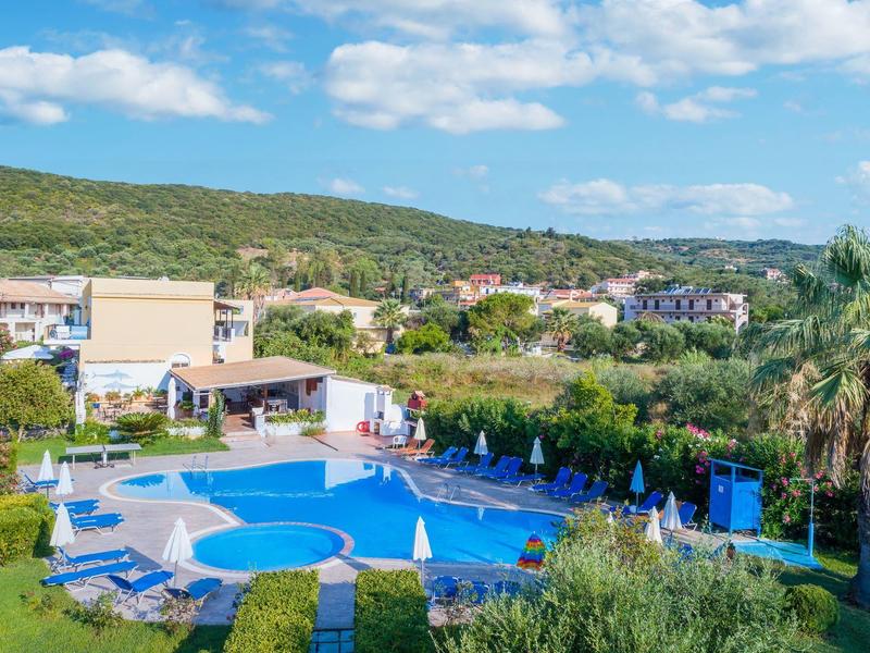 Outdoor hotel pool area with blue umbrellas, sunbeds, and surrounding greenery under a blue sky.