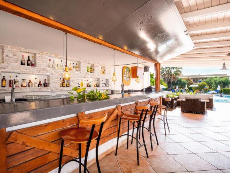 Outdoor bar with wooden counter and stools near a pool and lounge area under a covered roof.