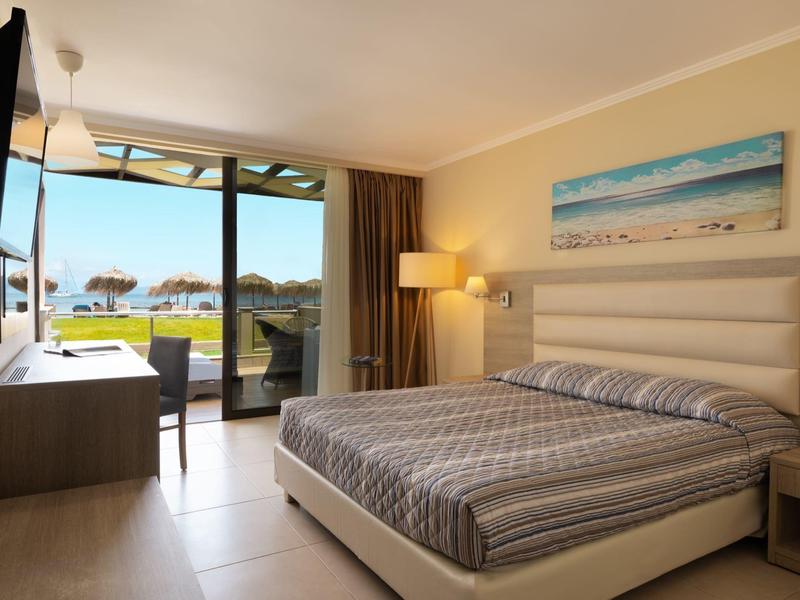 Chambre d'hôtel moderne avec lit double, télévision et vue sur la terrasse avec pelouse et parasols.