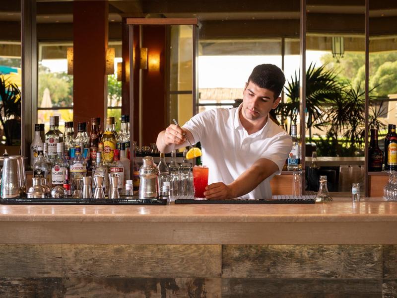 Barman prépare un cocktail dans un bar d'hôtel lumineux avec plusieurs bouteilles en arrière-plan.