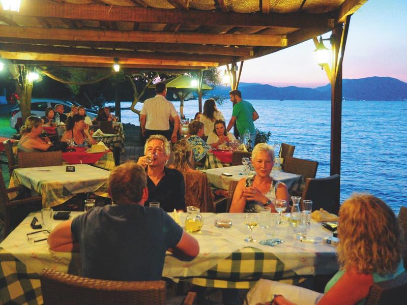 Menschen sitzen abends draußen in einem überdachten Strandrestaurant mit Blick aufs Meer.