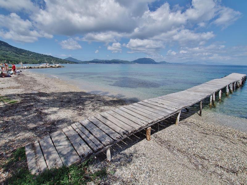 Holzerner Steg erstreckt sich über Kieselstrand ins blaue Meer unter hellem Himmel mit weißen Wolken.
