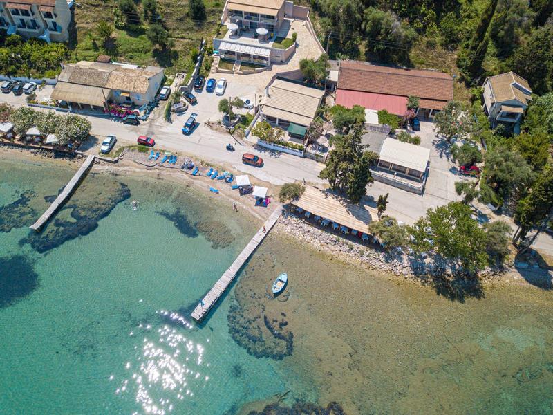 Kleiner Hafen mit Bootssteg, klarem Wasser, Badestrand und Gebäuden im grünen Umfeld.