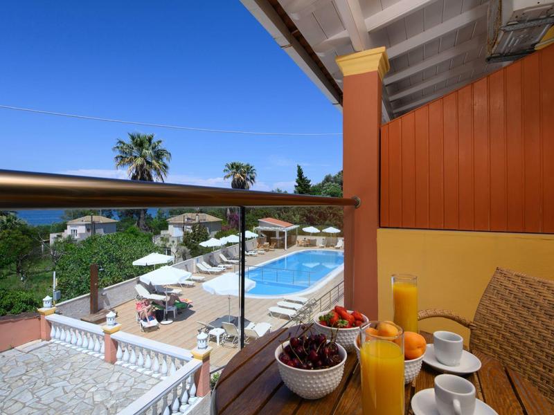 Balkon mit Frühstückstisch, Blick auf Pool, Palmen und blaue Himmel. Holzmöbel, Glasgeländer.