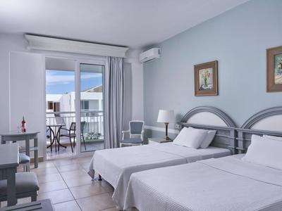 Bright hotel room with two beds, light blue walls, and a balcony with outdoor seating.