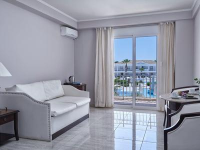 Modern hotel room with white sofa, armchair, and view of outdoor pool through glass doors.
