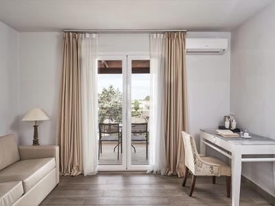 Bright hotel room with sofa, desk, chair, and balcony with curtains and air conditioner.