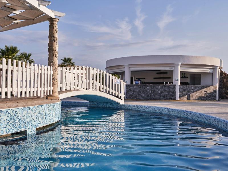 A curved pool with a white wooden bridge and a modern circular building in the background under blue sky.