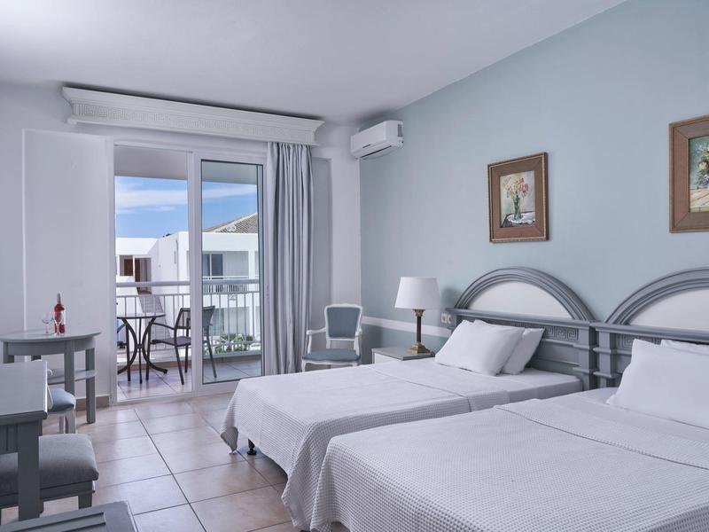 Bright hotel room with two beds, light blue walls, and a balcony with outdoor seating.