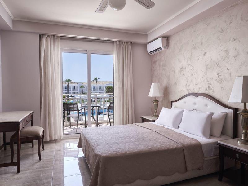 Bright hotel room with double bed, balcony overlooking buildings, desk, and lamps.