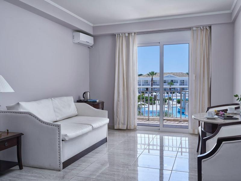Modern hotel room with white sofa, armchair, and view of outdoor pool through glass doors.