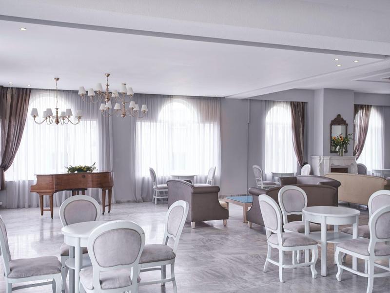 Bright hotel lounge with round tables, elegant chairs, chandeliers, and a grand piano near large windows.