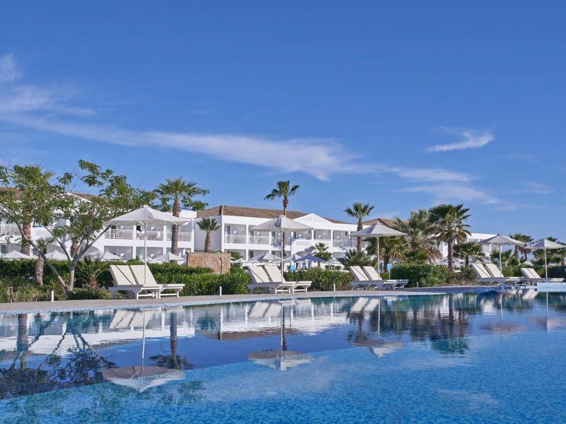 Resort with a large swimming pool, white buildings, palm trees, and clear blue sky.