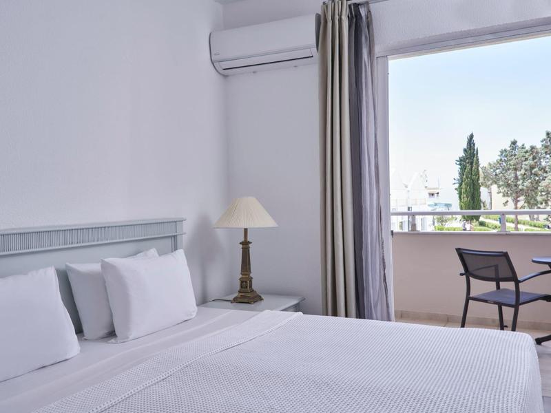 Bright hotel room with white bedding, bedside lamp, and balcony with table and chairs.