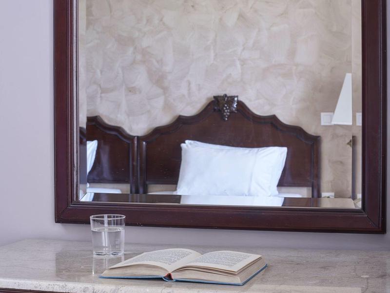 Wooden desk with an open book, glass of water, and mirror reflecting a bed with white pillow.