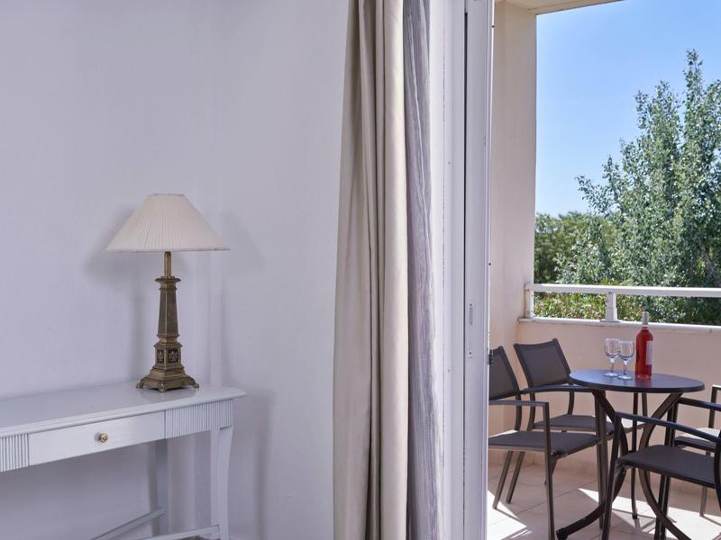 Balcony with table, chairs, and bottle, next to a lamp on a white desk inside a room.