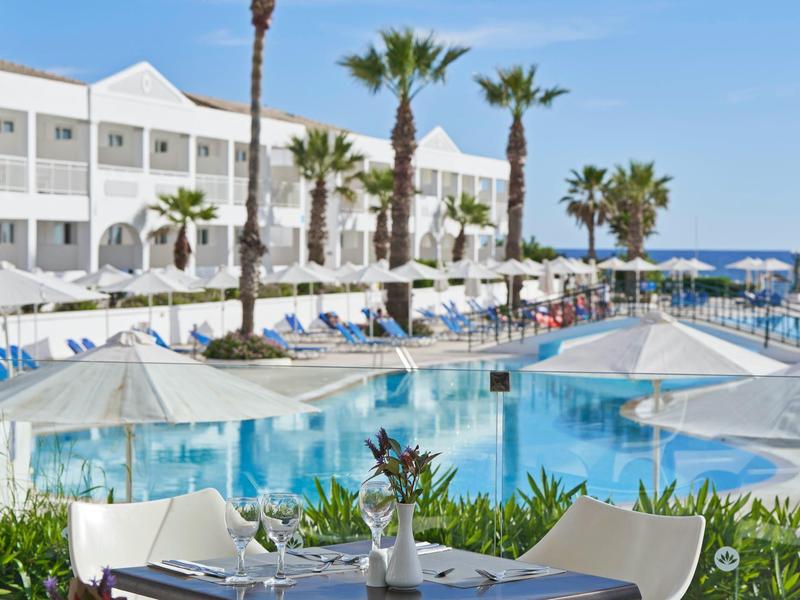 Outdoor dining area near a large pool with palm trees and white hotel building.