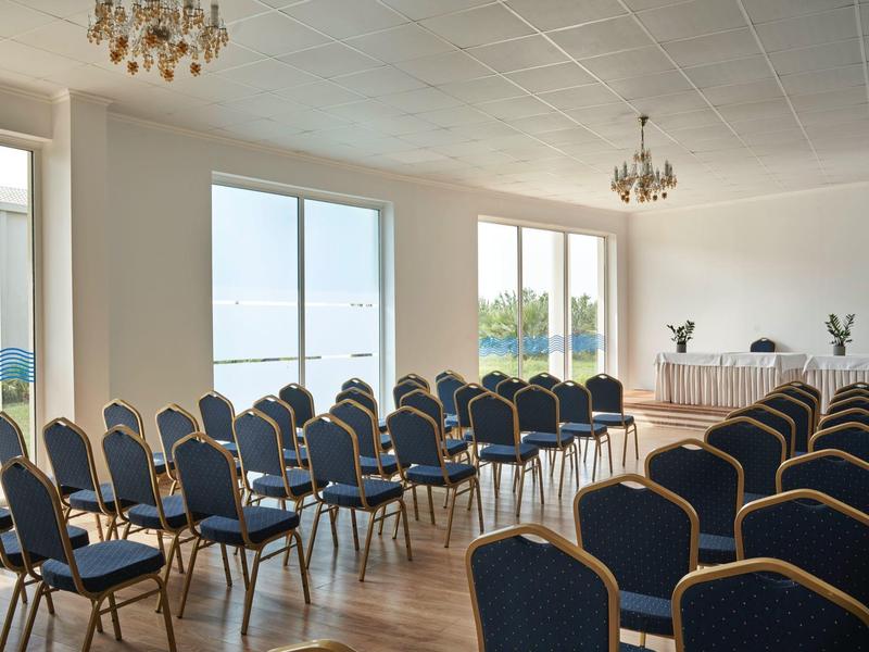 Bright conference room with rows of blue chairs and chandeliers near large windows.