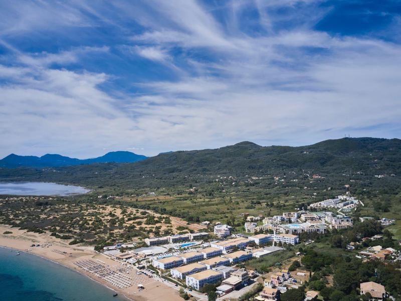 Coastal resort with sandy beach, white buildings, and green hills under a cloudy sky.