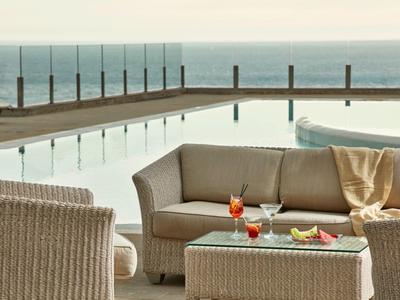 Outdoor-Lounge mit beigen Korbmöbeln, mehreren Getränken und Blick auf den Pool und das Meer im Hintergrund.