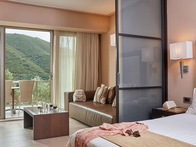 Modernes Hotelzimmer mit Bett, Sofa, Balkon und Blick auf grüne Hügel. Lichtdurchfluteter Raum.