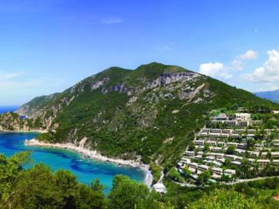 Berg mit grüner Vegetation, weißen Gebäuden an Hängen, blauem Meer und wolkigem Himmel.