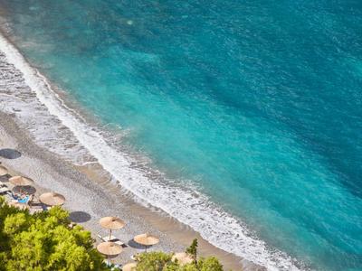 Strand mit Kies, türkisblauem Meer und Sonnenschirmen, grüne Pflanzen im Vordergrund.
