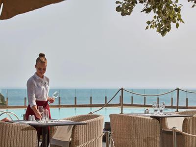 Eine Kellnerin serviert Getränke auf einer Terrasse mit Blick auf das Meer und den Strand.