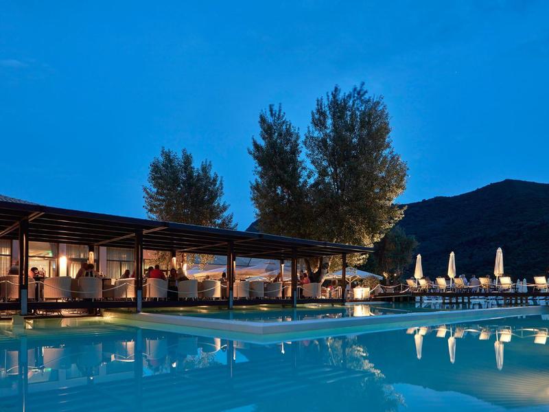 Schwimmbad bei Dämmerung mit beleuchtetem Pavillon, Bäumen und Bergen im Hintergrund.