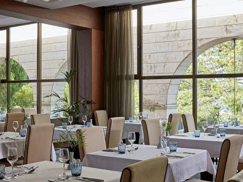 Modernes Restaurant mit weißen Tischdecken, großen Fenstern und Blick auf Garten mit Steinbogen.