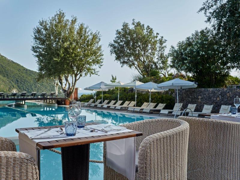 Luxuriöse Outdoor-Sitzplätze am Pool mit Tisch, Stühlen, Sonnenschirmen und grüner Landschaft im Hintergrund.