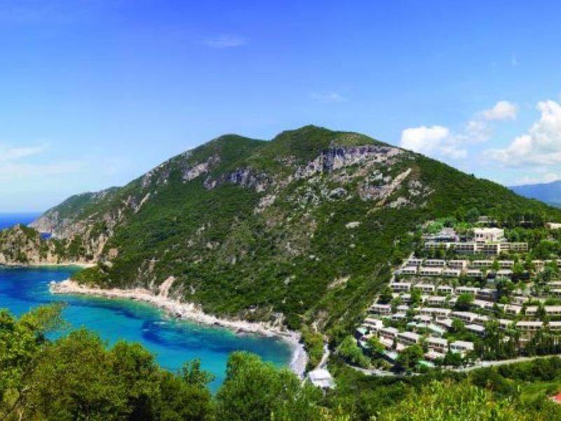 Berg mit grüner Vegetation, weißen Gebäuden an Hängen, blauem Meer und wolkigem Himmel.
