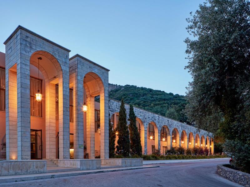 Lange Steinfassade mit hohen Bögen und warmem Licht vor bewaldetem Hügel bei blauem Himmel.