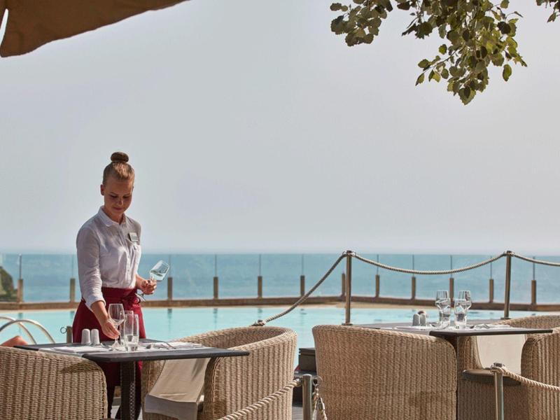 Eine Kellnerin trägt ein Tablett am Strandrestaurant mit Blick aufs blaue Meer bei klarem Himmel.