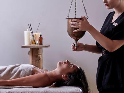 Frau liegt entspannt, während Therapeutin warmes Öl aus Gefäß auf Stirn gießt, ruhiger Raum mit Kerzen.