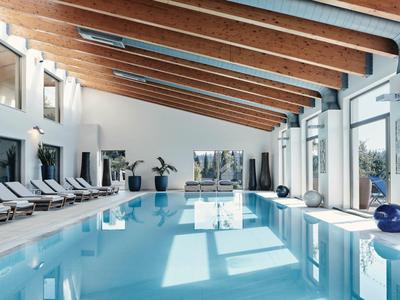 Ein heller Innenpoolraum mit Holzdeckenbalken, großen Fenstern und Liegestühlen am Beckenrand.