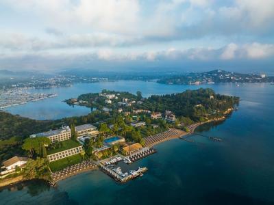 Widok z lotu ptaka na nadmorski kompleks z hotelem, plażą i zalesionymi wzgórzami pod zachmurzonym niebem.
