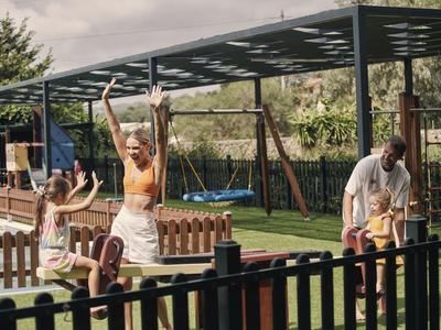 Dzieci bawią się na huśtawce z dorosłym w tle w ogrodzie z ogrodzeniem.