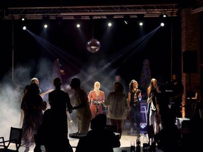 Ludzie stojący na scenie z reflektorami i mgłą w tle.