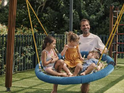 Ein Mann schaukelt mit drei Kindern auf einem großen blauen Nestschaukel im Garten.
